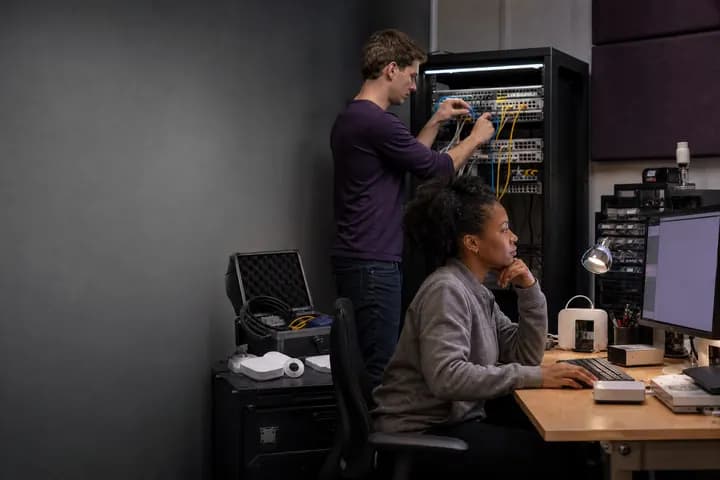 Two engineers working at a server rack and workstation in a technology company office
